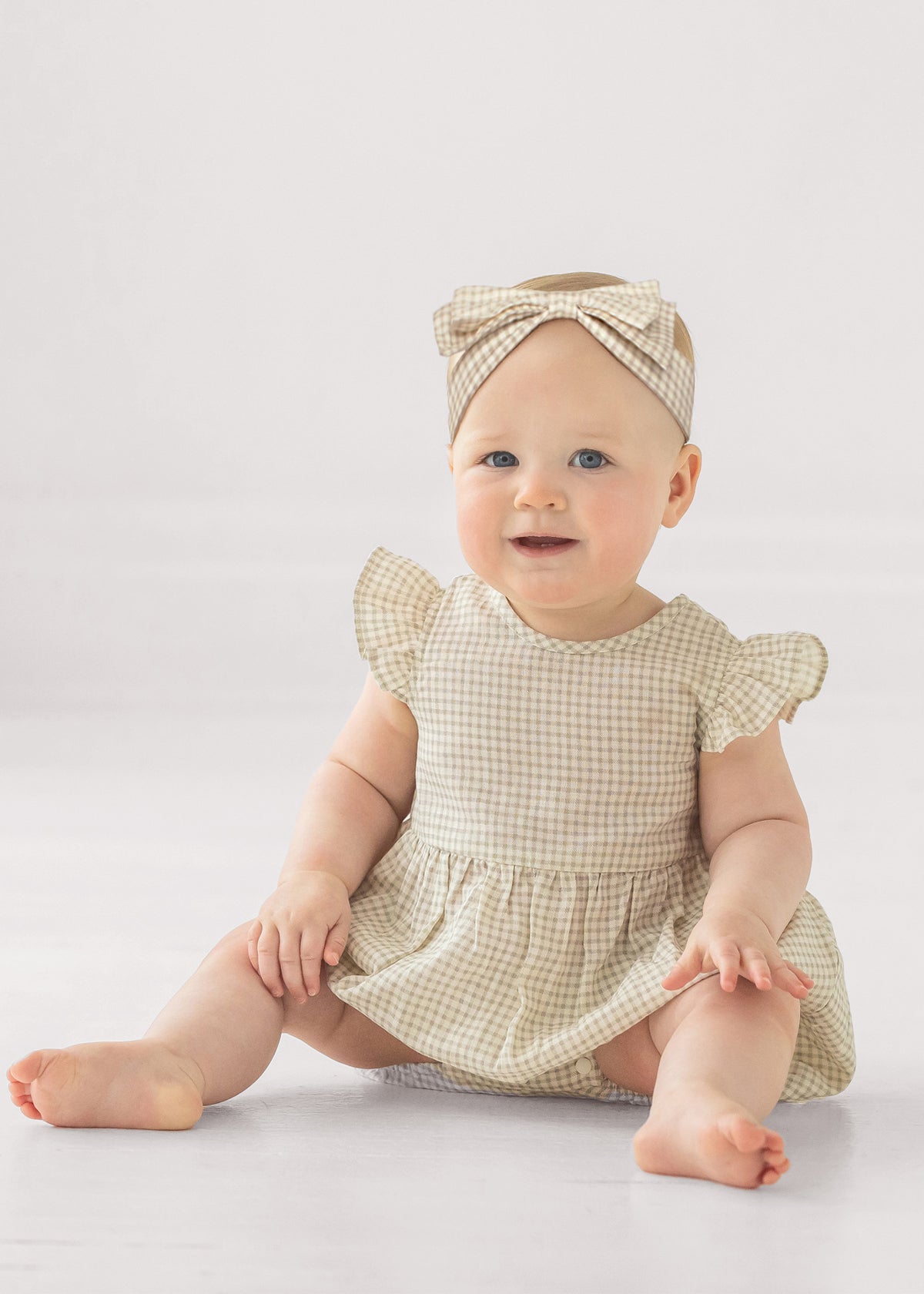 A baby in the Mabel and Honey Dakota Ruffled Short Sleeve Romper, featuring a beige and white checkered pattern with ruffled sleeves and a matching headband, sits smiling on the floor against a plain light background.
