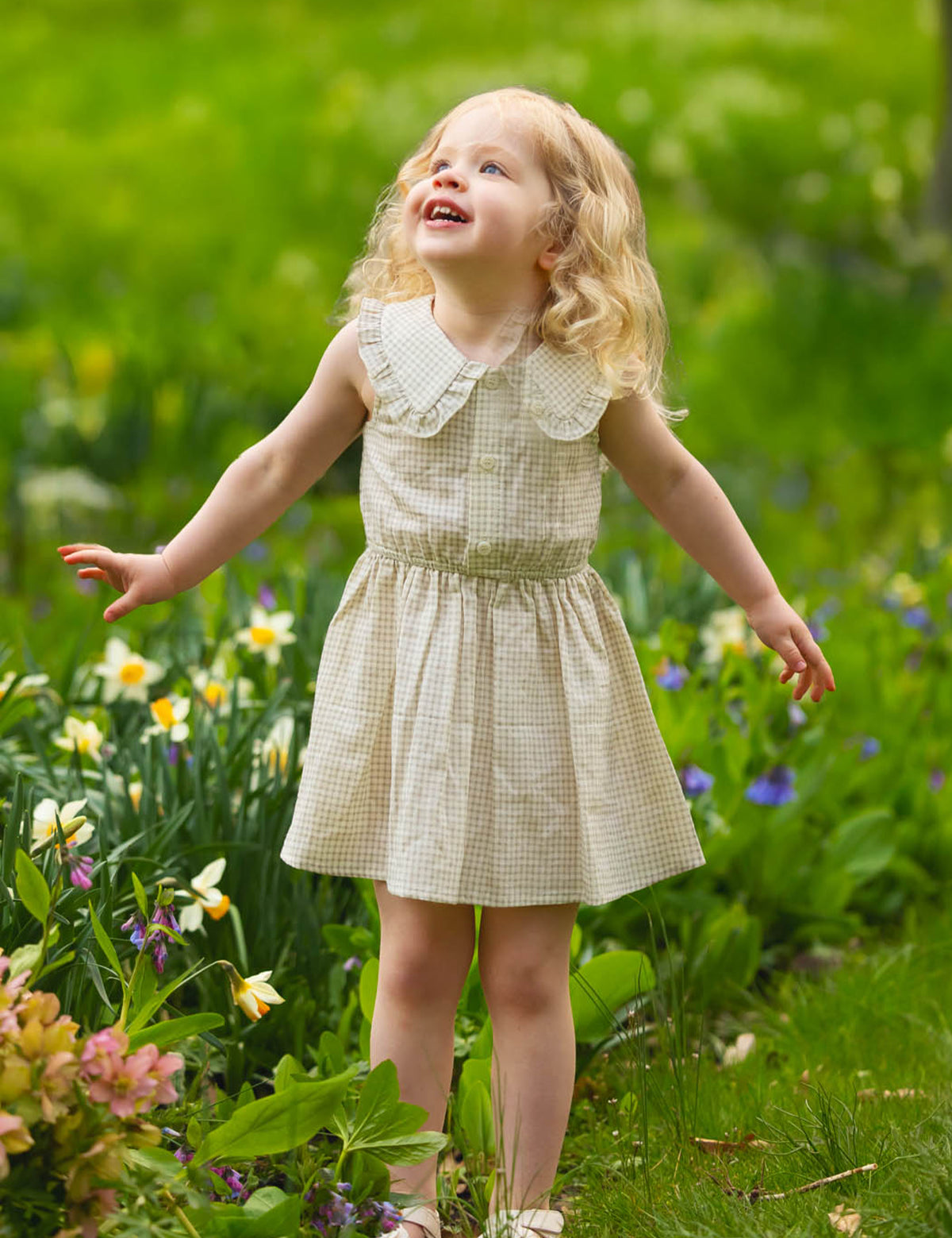 A young girl with blonde curls wears the Mabel and Honey Dakota Sleeveless Dress as she stands outdoors among blooming flowers, smiling and looking up with her arms slightly outstretched.