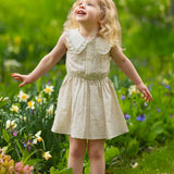 A young girl with blonde curls wears the Mabel and Honey Dakota Sleeveless Dress as she stands outdoors among blooming flowers, smiling and looking up with her arms slightly outstretched.