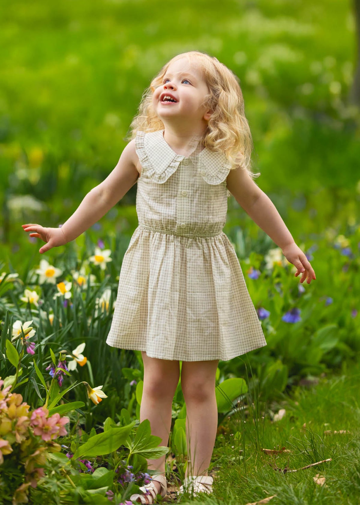 A young girl with blonde curls wears the Mabel and Honey Dakota Sleeveless Dress as she stands outdoors among blooming flowers, smiling and looking up with her arms slightly outstretched.