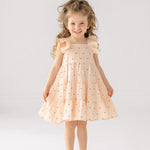 A young girl with curly hair smiles in the Mabel and Honey Stargirl Ruffled Sleeve Dress—a light pink, tiered cotton dress with black stars—paired with sandals, standing against a plain white background.
