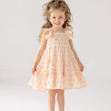 A young girl with curly hair smiles in the Mabel and Honey Stargirl Ruffled Sleeve Dress—a light pink, tiered cotton dress with black stars—paired with sandals, standing against a plain white background.