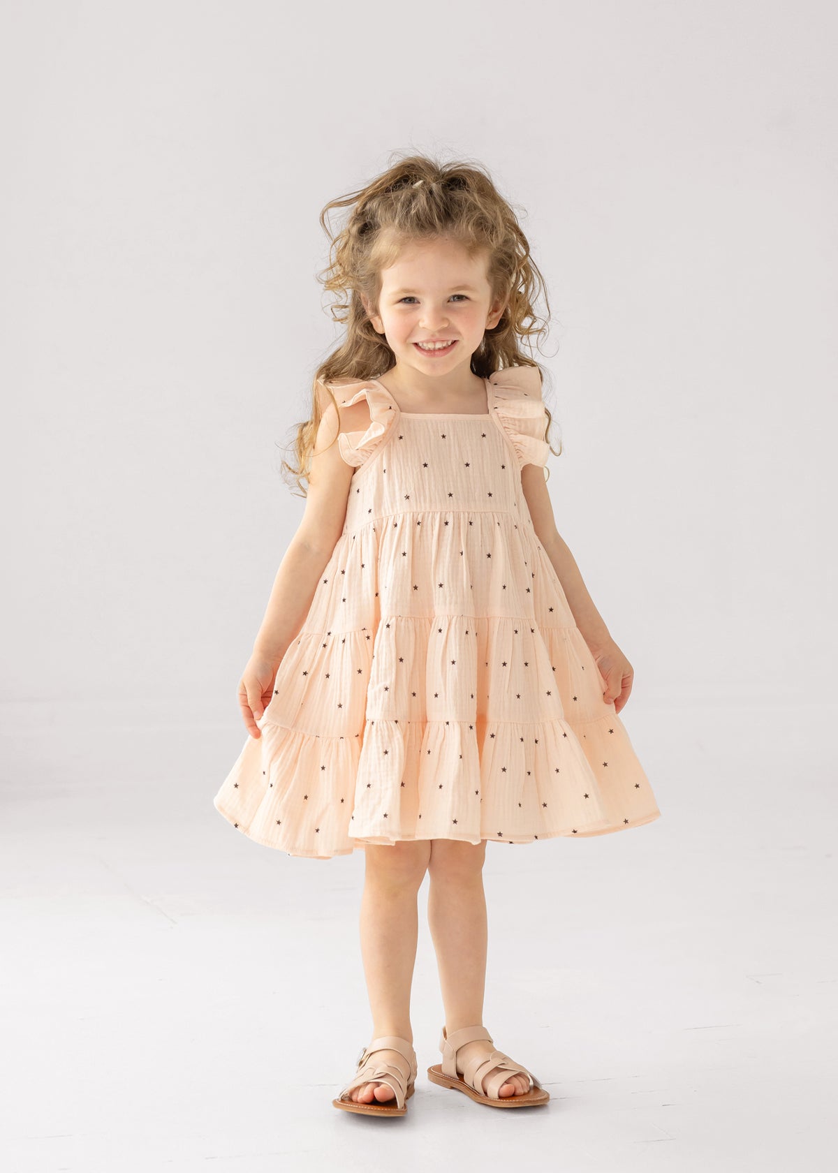 A young girl with curly hair smiles in the Mabel and Honey Stargirl Ruffled Sleeve Dress—a light pink, tiered cotton dress with black stars—paired with sandals, standing against a plain white background.