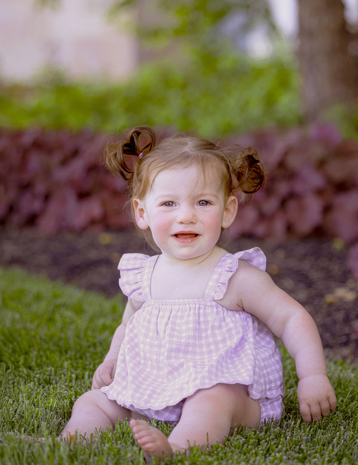 A young toddler with light skin and brown hair in pigtails smiles barefoot on green grass, wearing the Sugar Plum Sleeveless Romper by Mabel and Honey, surrounded by plants and trees outdoors.