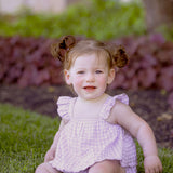A young toddler with light skin and brown hair in pigtails smiles barefoot on green grass, wearing the Sugar Plum Sleeveless Romper by Mabel and Honey, surrounded by plants and trees outdoors.