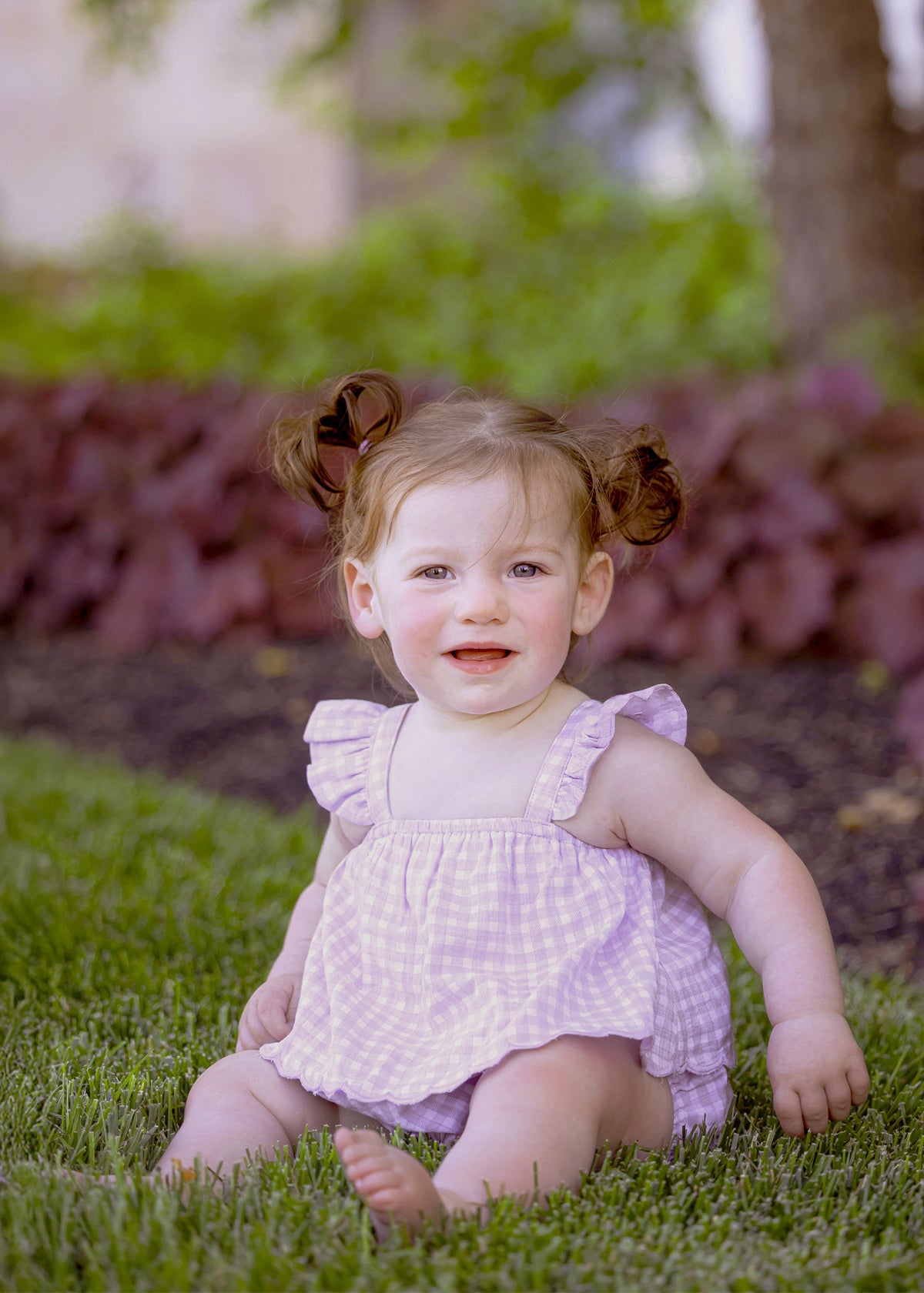 A young toddler with light skin and brown hair in pigtails smiles barefoot on green grass, wearing the Sugar Plum Sleeveless Romper by Mabel and Honey, surrounded by plants and trees outdoors.