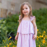 A young girl with light brown hair smiles softly outdoors, wearing the Mabel and Honey Sugar Plum Sleeveless Dress—a pink and white gingham style with ruffled straps—standing in front of green bushes and yellow flowers.