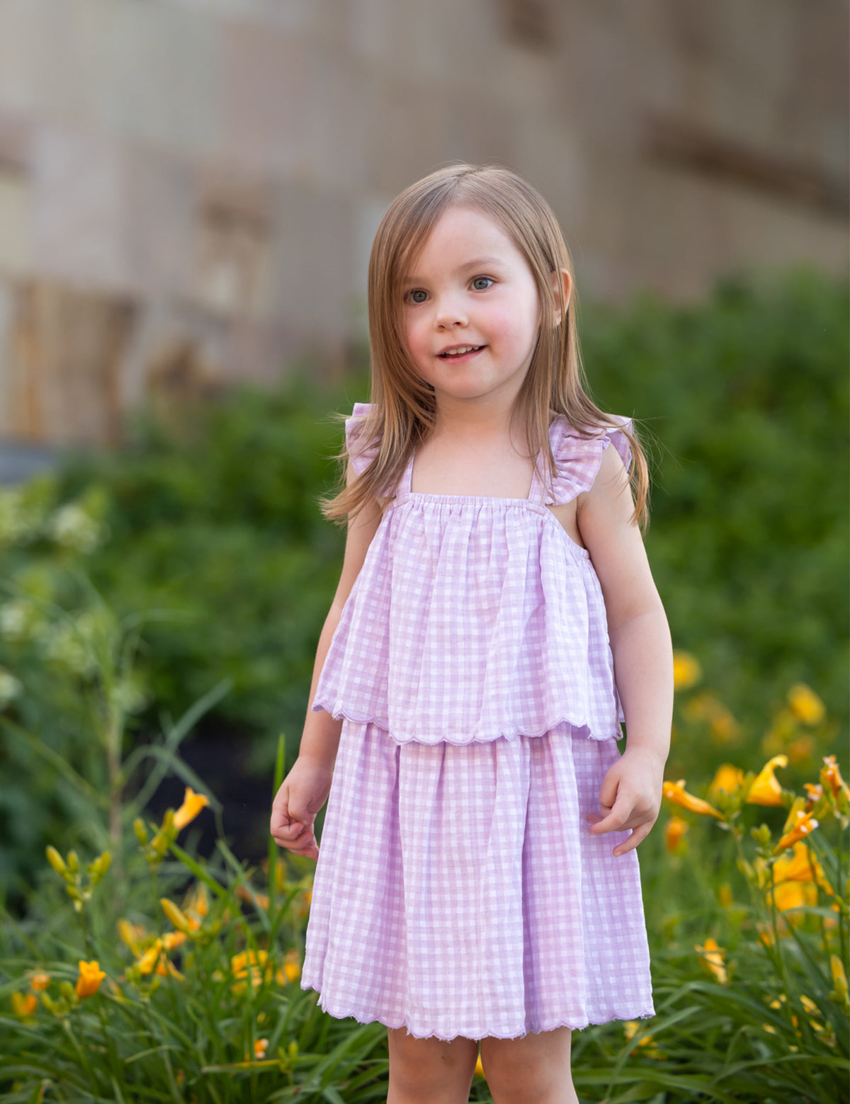 A young girl with light brown hair smiles softly outdoors, wearing the Mabel and Honey Sugar Plum Sleeveless Dress—a pink and white gingham style with ruffled straps—standing in front of green bushes and yellow flowers.