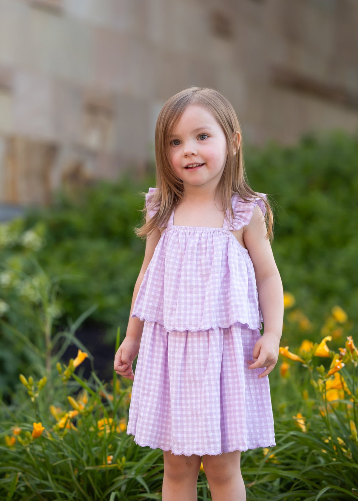 A young girl with light brown hair smiles softly outdoors, wearing the Mabel and Honey Sugar Plum Sleeveless Dress—a pink and white gingham style with ruffled straps—standing in front of green bushes and yellow flowers.