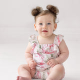 A smiling baby with brown hair in two buns and blue eyes sits on a white surface, wearing the Rosie Ribbons Ruffled Tank Romper by Mabel and Honey, featuring floral pink and green patterns with ruffled shoulder straps.