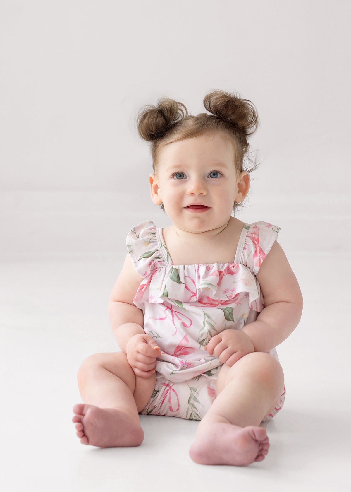 A smiling baby with brown hair in two buns and blue eyes sits on a white surface, wearing the Rosie Ribbons Ruffled Tank Romper by Mabel and Honey, featuring floral pink and green patterns with ruffled shoulder straps.