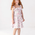 A young girl with long red hair smiles as she stands against a white background, wearing the Rosie Ribbons Ruffled Tank Dress by Mabel and Honey paired with white sandals.