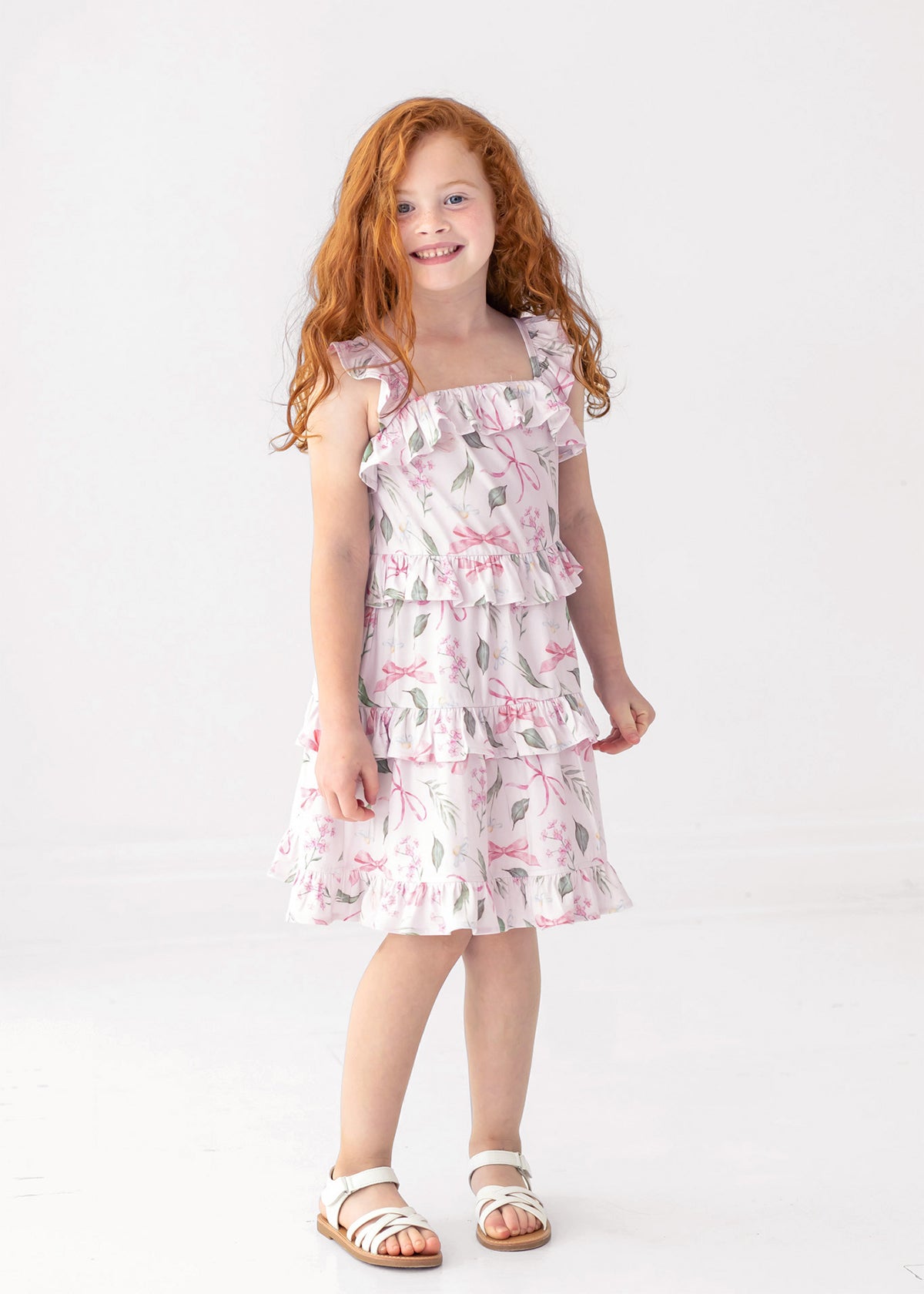 A young girl with long red hair smiles as she stands against a white background, wearing the Rosie Ribbons Ruffled Tank Dress by Mabel and Honey paired with white sandals.