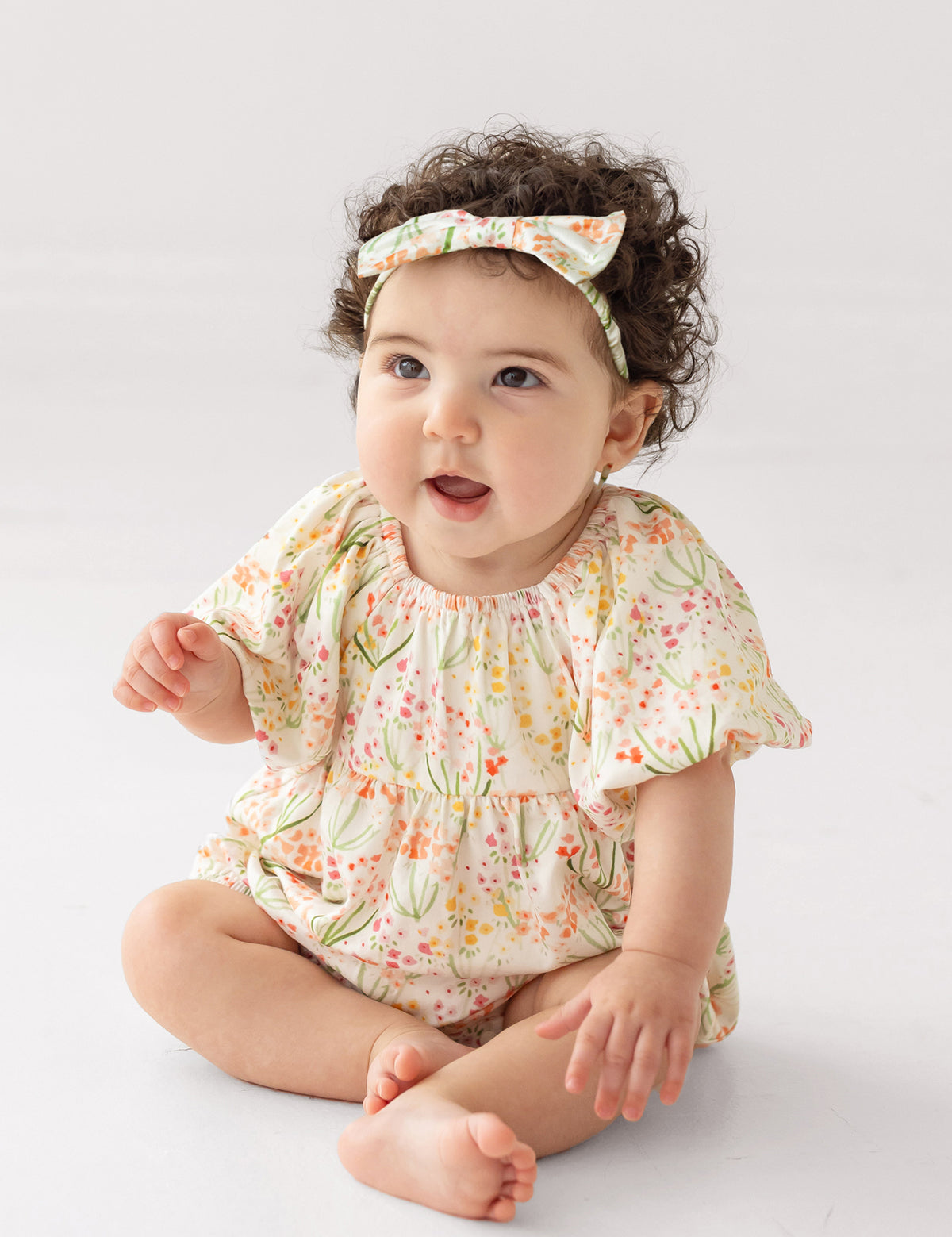 A baby with curly dark hair, wearing Mabel and Honey’s Springtime Garden Short Puff Sleeve Romper and a matching headband, sits barefoot on a light background, gazing upward with an open-mouthed smile.