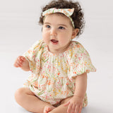 A baby with curly dark hair, wearing Mabel and Honey’s Springtime Garden Short Puff Sleeve Romper and a matching headband, sits barefoot on a light background, gazing upward with an open-mouthed smile.