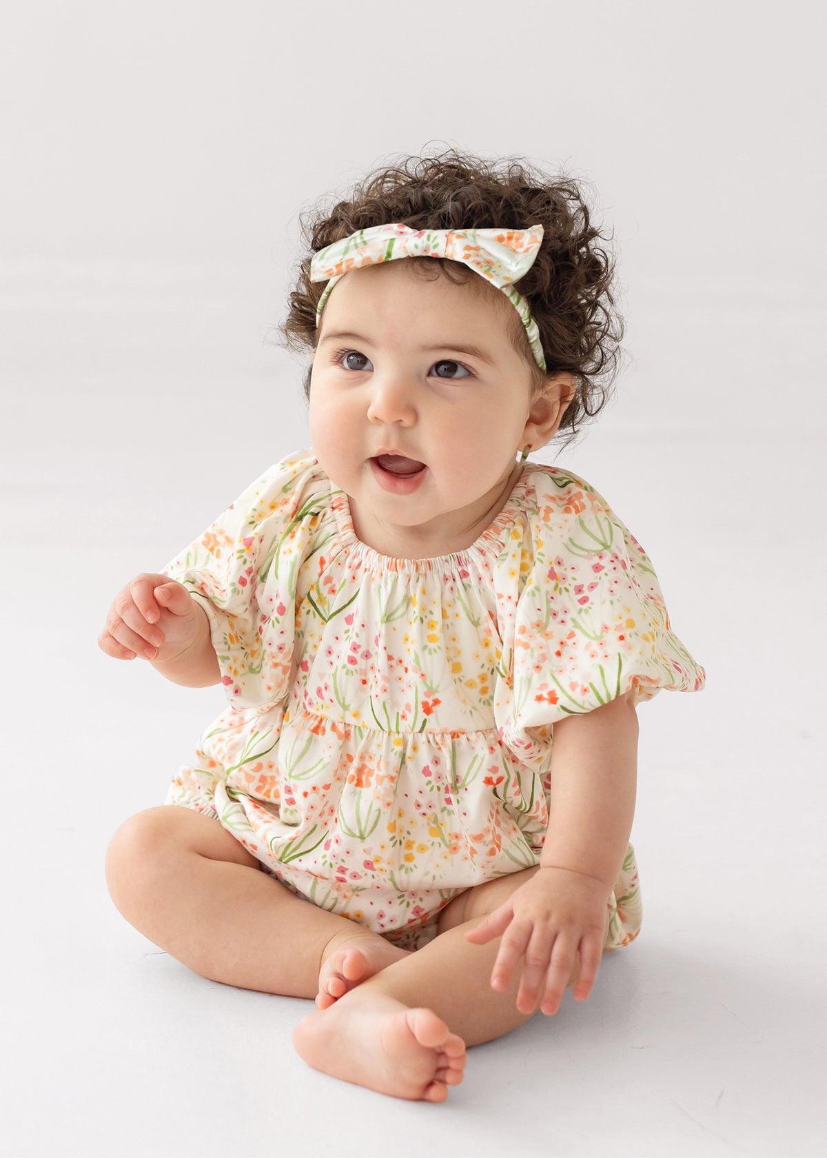 A baby with curly dark hair, wearing Mabel and Honey’s Springtime Garden Short Puff Sleeve Romper and a matching headband, sits barefoot on a light background, gazing upward with an open-mouthed smile.