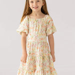 A young girl with long light brown hair smiles in the Mabel and Honey Springtime Garden Short Puff Sleeve Dress, featuring colorful floral prints. She stands against a plain light background.