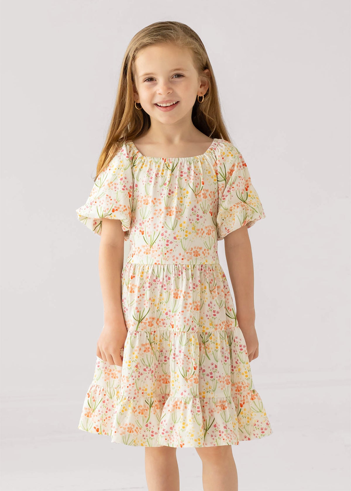 A young girl with long light brown hair smiles in the Mabel and Honey Springtime Garden Short Puff Sleeve Dress, featuring colorful floral prints. She stands against a plain light background.