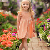 A young girl smiles on a garden path in the Mabel and Honey Sunkissed Apricot Short Flutter Sleeve Dress, featuring peach stripes. She has long blonde hair and white sandals, surrounded by vibrant flowers and greenery.