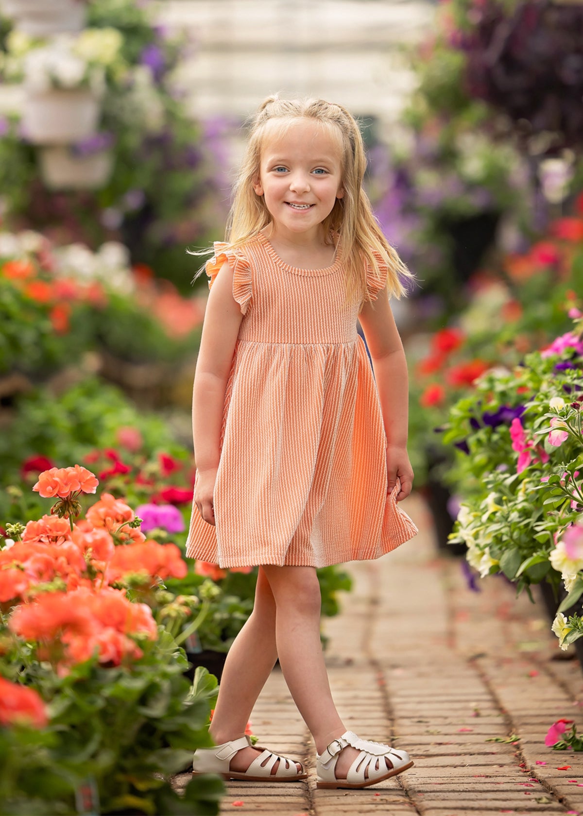 A young girl smiles on a garden path in the Mabel and Honey Sunkissed Apricot Short Flutter Sleeve Dress, featuring peach stripes. She has long blonde hair and white sandals, surrounded by vibrant flowers and greenery.