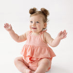 A smiling baby with light hair in two small buns wears the Pinky Linky Sleeveless with Knot Detail Romper by Mabel and Honey, sitting on the floor with arms raised against a plain white background.