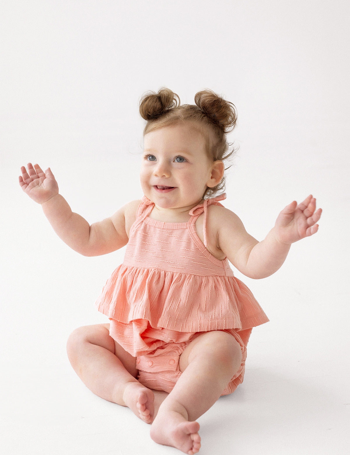 A smiling baby with light hair in two small buns wears the Pinky Linky Sleeveless with Knot Detail Romper by Mabel and Honey, sitting on the floor with arms raised against a plain white background.