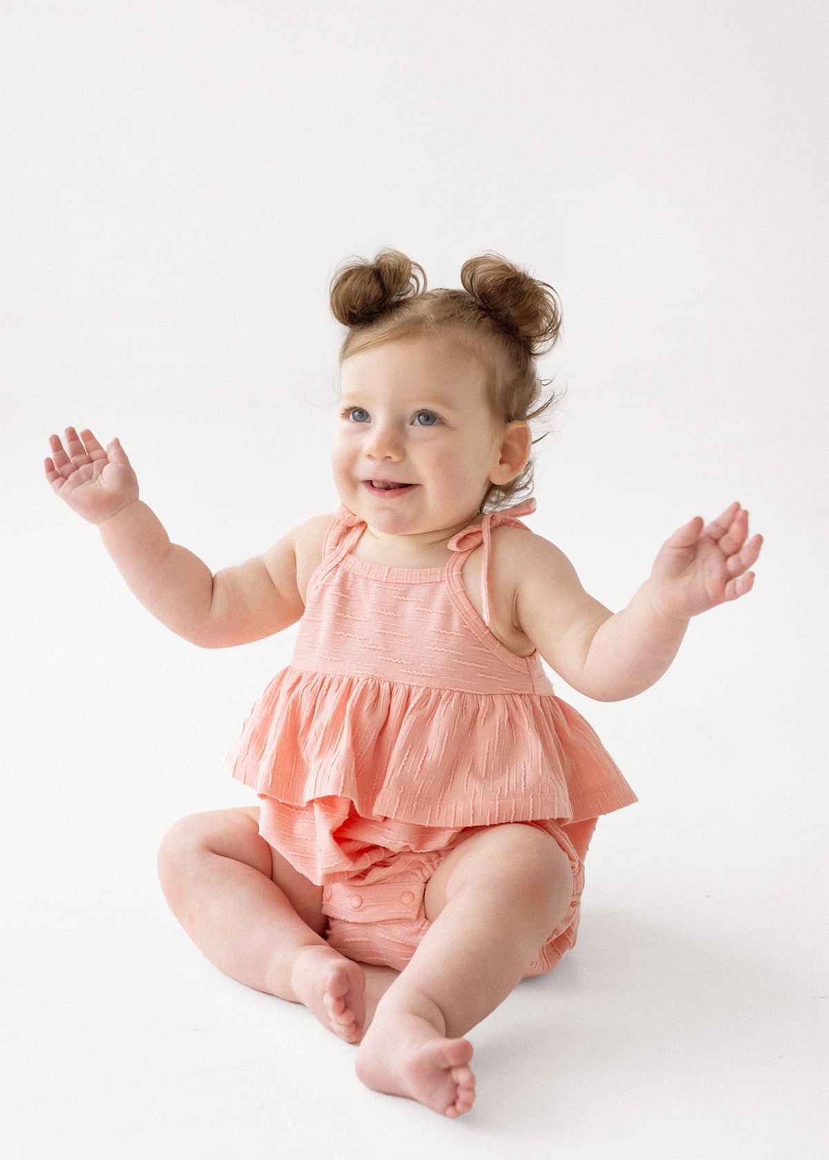 A smiling baby with light hair in two small buns wears the Pinky Linky Sleeveless with Knot Detail Romper by Mabel and Honey, sitting on the floor with arms raised against a plain white background.
