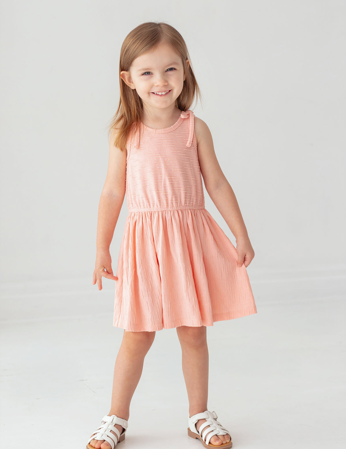 A young girl with light brown hair smiles against a white background, wearing the Mabel and Honey Pinky Linky Sleeveless with Knot Detail Dress and white sandals.