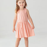A young girl with light brown hair smiles against a white background, wearing the Mabel and Honey Pinky Linky Sleeveless with Knot Detail Dress and white sandals.