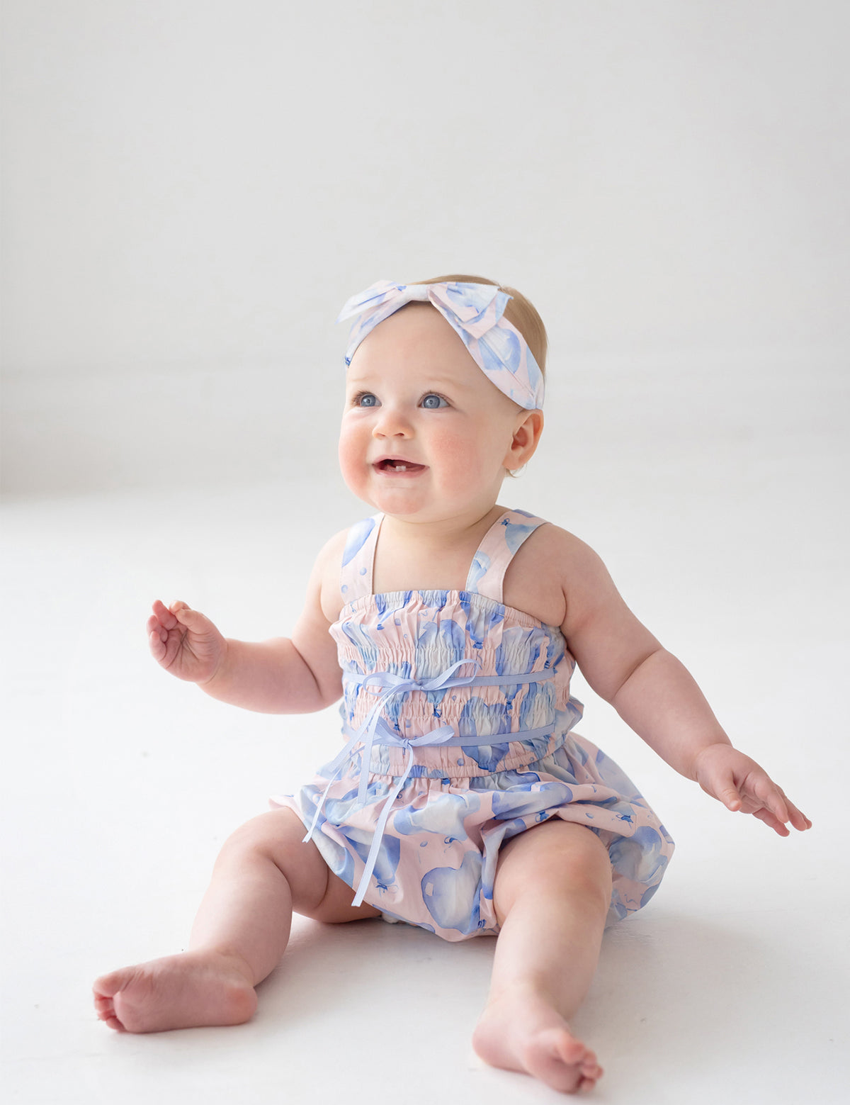 A baby in the Mabel and Honey Up and Away Sleeveless Romper, pastel blue with hearts, sits on a white floor wearing a matching headband, smiling upward with arms raised and legs stretched out.