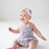 A baby in the Mabel and Honey Up and Away Sleeveless Romper, pastel blue with hearts, sits on a white floor wearing a matching headband, smiling upward with arms raised and legs stretched out.
