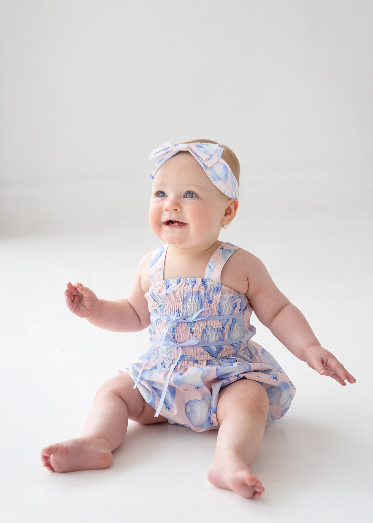 A baby in the Mabel and Honey Up and Away Sleeveless Romper, pastel blue with hearts, sits on a white floor wearing a matching headband, smiling upward with arms raised and legs stretched out.