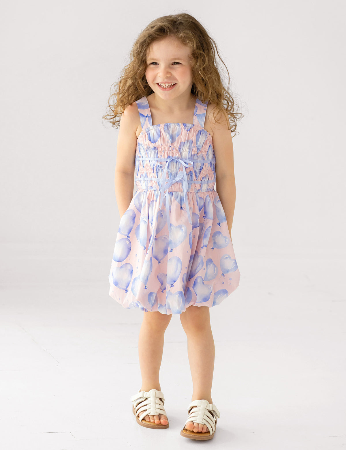 A young girl with long curly hair smiles with her hands in her pockets, wearing the Mabel and Honey "Up and Away" Sleeveless Dress in a pastel pink and blue balloon print, paired with white sandals, against a plain white background.
