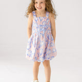 A young girl with long curly hair smiles with her hands in her pockets, wearing the Mabel and Honey "Up and Away" Sleeveless Dress in a pastel pink and blue balloon print, paired with white sandals, against a plain white background.
