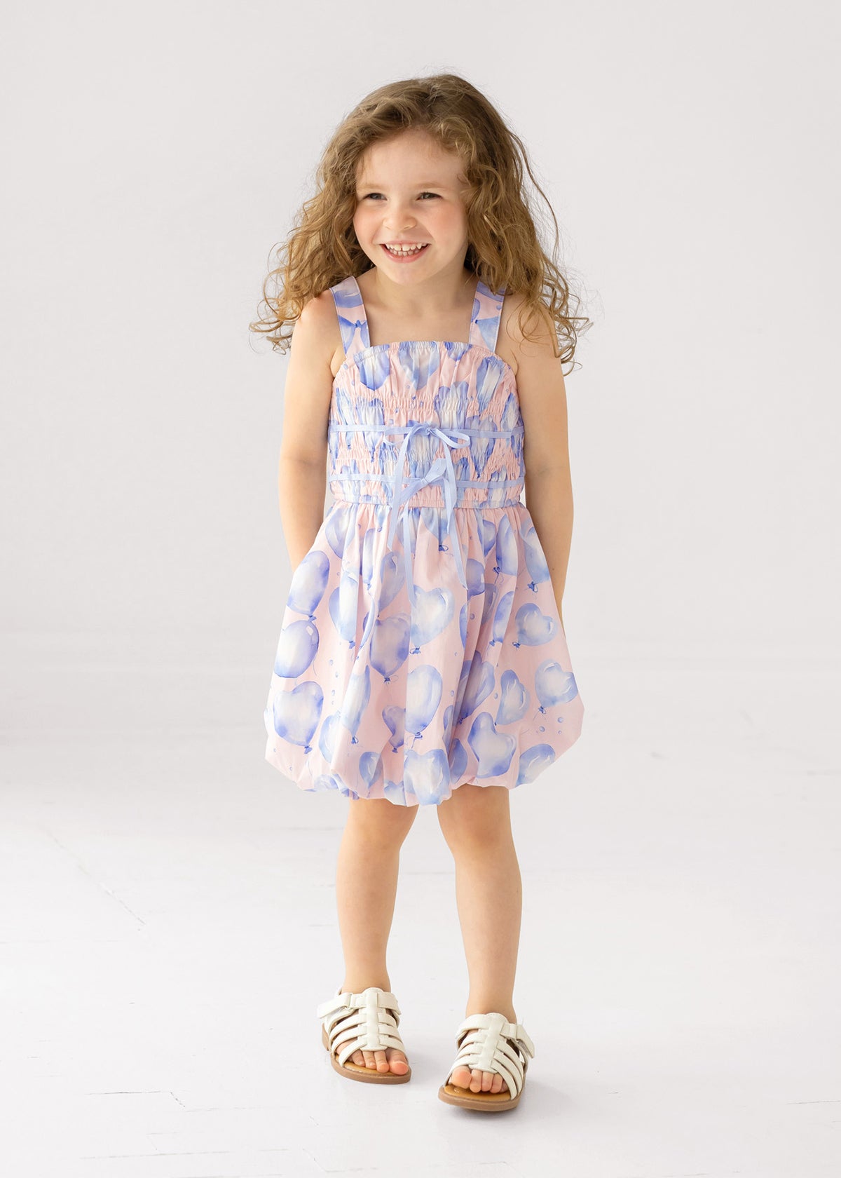 A young girl with long curly hair smiles with her hands in her pockets, wearing the Mabel and Honey "Up and Away" Sleeveless Dress in a pastel pink and blue balloon print, paired with white sandals, against a plain white background.