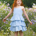 A young girl with long red hair smiles in a field of wildflowers, wearing the Mabel and Honey Up and Away Sleeveless Tiered Dress, with green trees in the background.