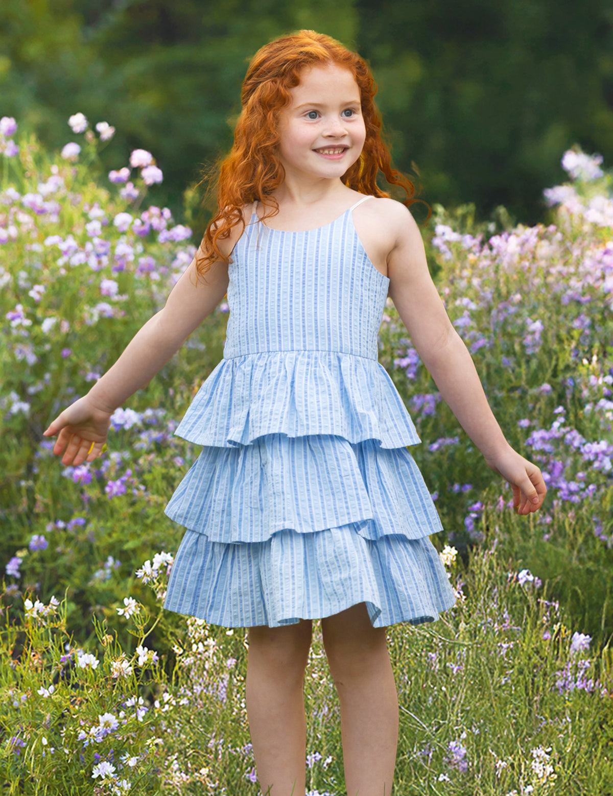 A young girl with long red hair smiles in a field of wildflowers, wearing the Mabel and Honey Up and Away Sleeveless Tiered Dress, with green trees in the background.