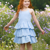 A young girl with long red hair smiles in a field of wildflowers, wearing the Mabel and Honey Up and Away Sleeveless Tiered Dress, with green trees in the background.