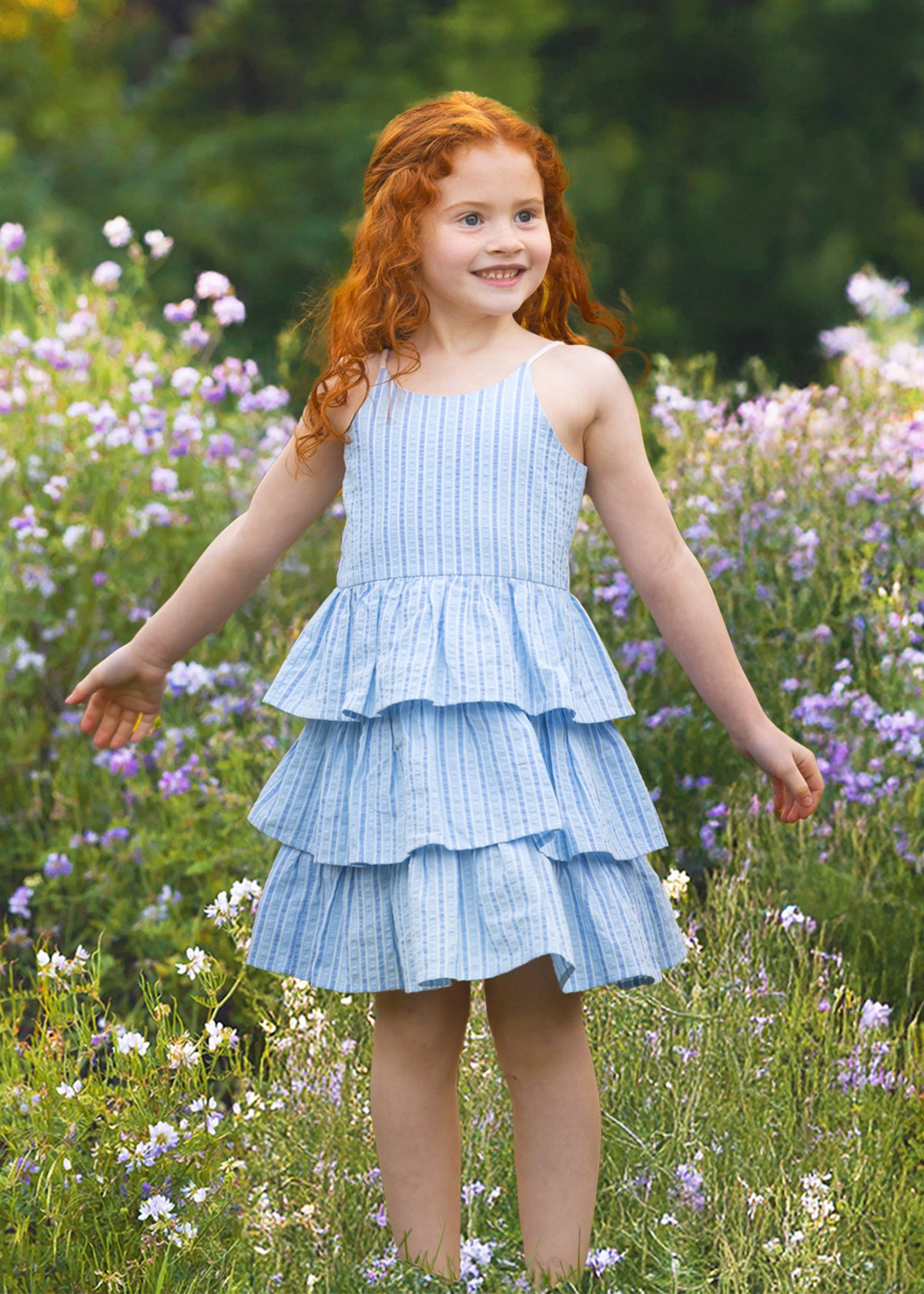 A young girl with long red hair smiles in a field of wildflowers, wearing the Mabel and Honey Up and Away Sleeveless Tiered Dress, with green trees in the background.
