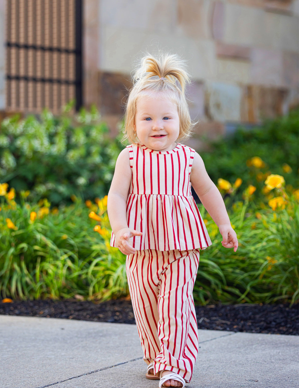 A young toddler with blond hair smiles while walking outdoors in the Mabel and Honey Terracotta Stripe Sleeveless Two Piece Set, surrounded by green plants and yellow flowers.