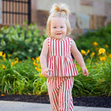 A young toddler with blond hair smiles while walking outdoors in the Mabel and Honey Terracotta Stripe Sleeveless Two Piece Set, surrounded by green plants and yellow flowers.