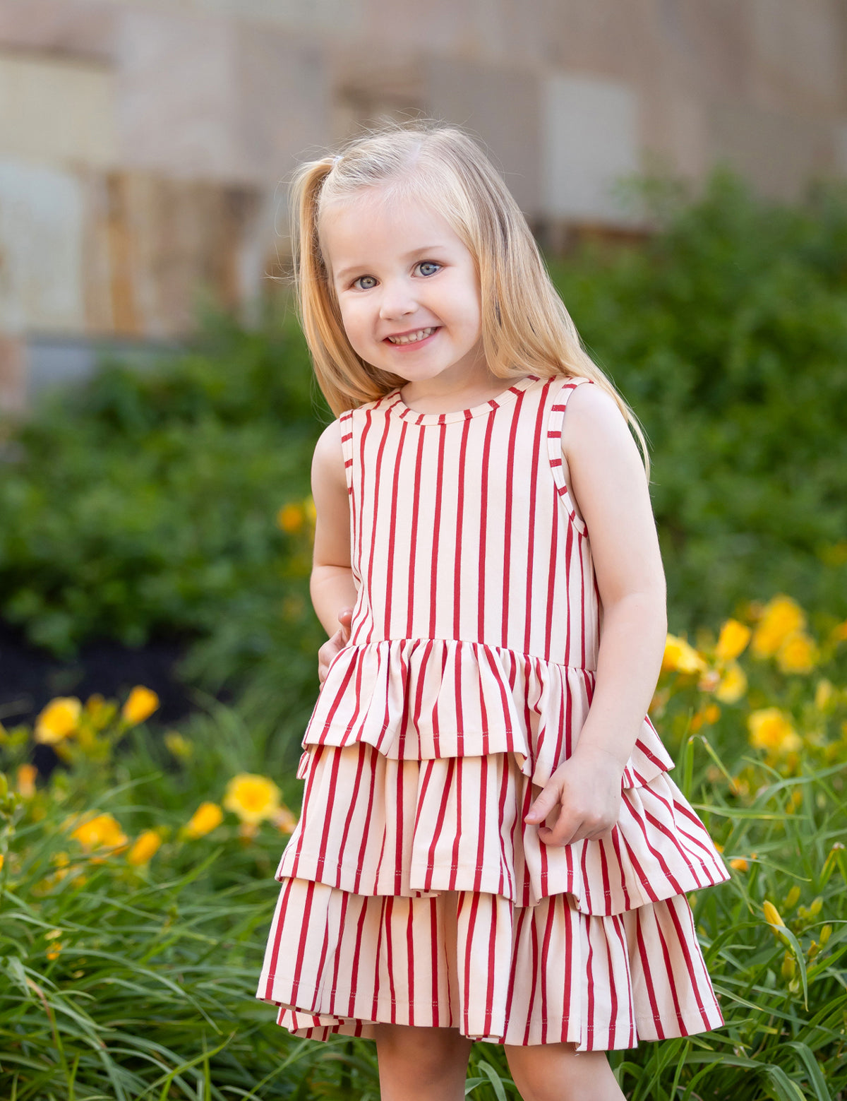 A young girl with long blonde hair smiles in front of green plants and yellow flowers, wearing the Mabel and Honey Terracotta Stripe Sleeveless Dress with a three-tiered skirt.