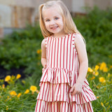 A young girl with long blonde hair smiles in front of green plants and yellow flowers, wearing the Mabel and Honey Terracotta Stripe Sleeveless Dress with a three-tiered skirt.