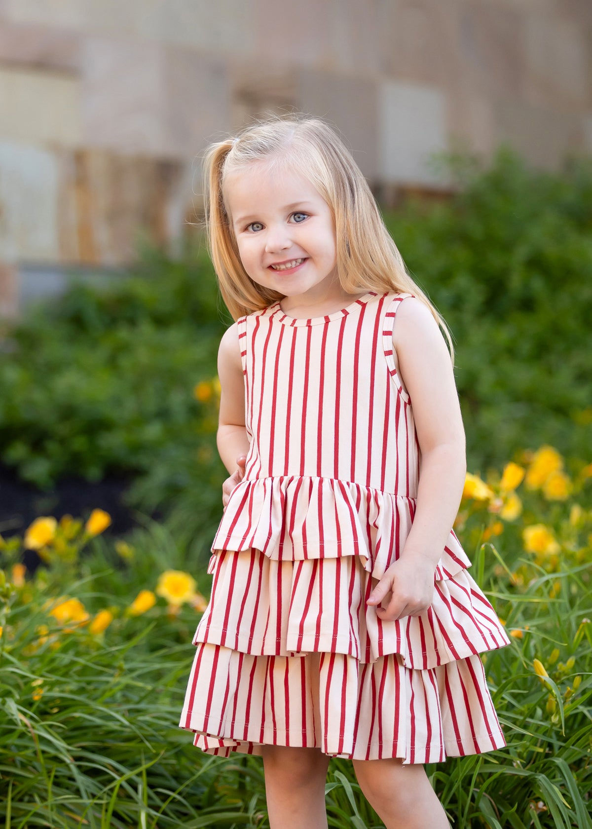 A young girl with long blonde hair smiles in front of green plants and yellow flowers, wearing the Mabel and Honey Terracotta Stripe Sleeveless Dress with a three-tiered skirt.