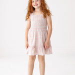 A young girl with long red curly hair smiles while wearing the Sawyer Sleeveless Dress by Mabel and Honey, featuring a smocked bodice and ruffled hem, paired with white sandals against a plain white background.