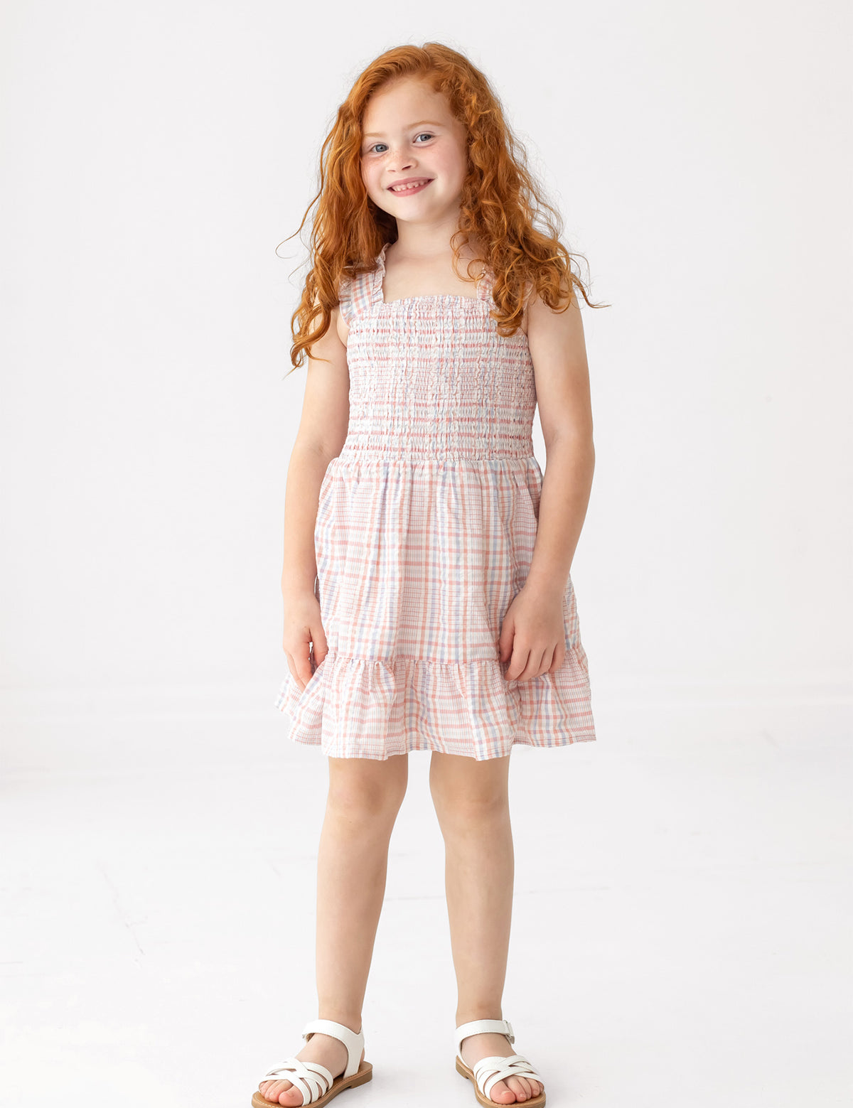 A young girl with long red curly hair smiles while wearing the Sawyer Sleeveless Dress by Mabel and Honey, featuring a smocked bodice and ruffled hem, paired with white sandals against a plain white background.