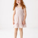 A young girl with long red curly hair smiles while wearing the Sawyer Sleeveless Dress by Mabel and Honey, featuring a smocked bodice and ruffled hem, paired with white sandals against a plain white background.