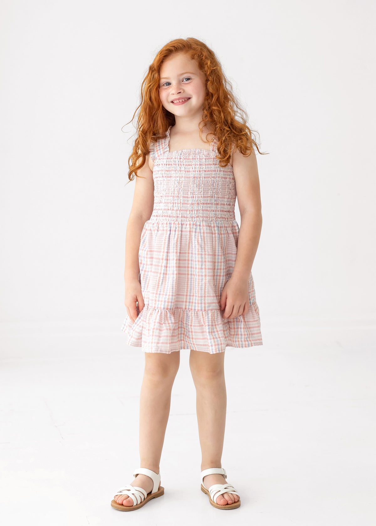 A young girl with long red curly hair smiles while wearing the Sawyer Sleeveless Dress by Mabel and Honey, featuring a smocked bodice and ruffled hem, paired with white sandals against a plain white background.