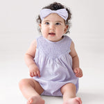 A baby with curly hair and a light purple headband wears the Magnolia Sleeveless Romper by Mabel and Honey, sitting on a white floor and gazing upward with a curious expression.