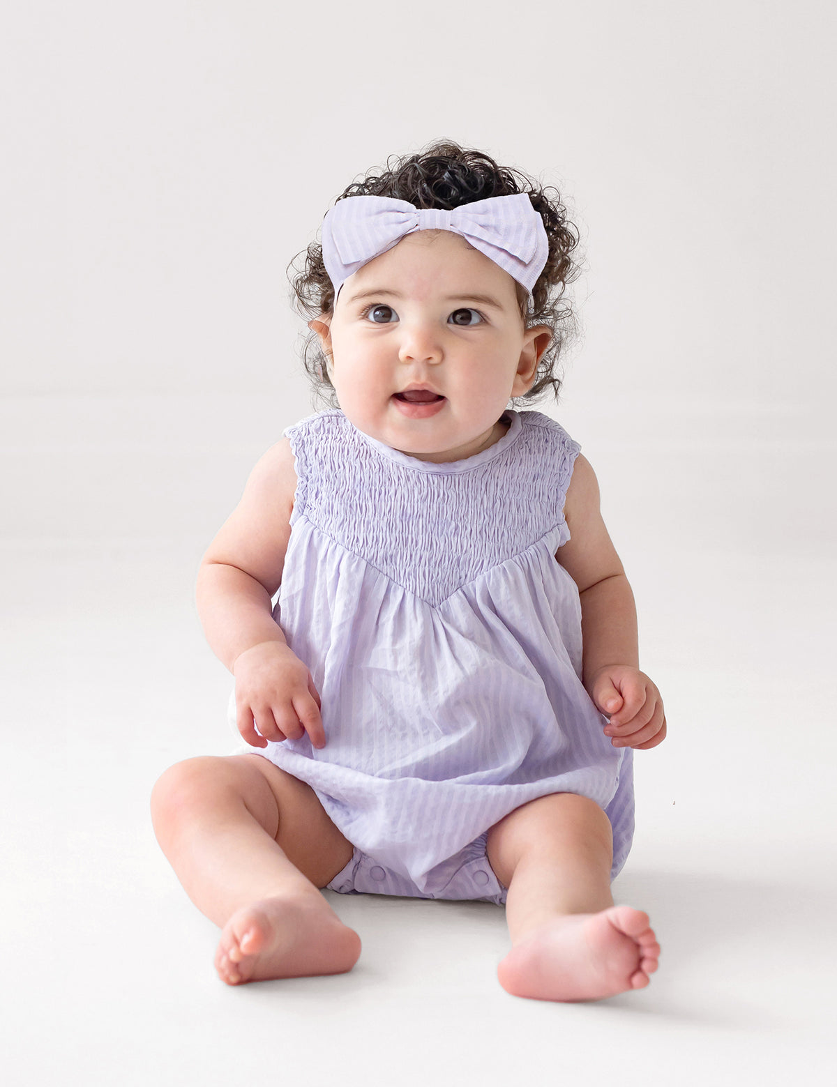 A baby with curly hair and a light purple headband wears the Magnolia Sleeveless Romper by Mabel and Honey, sitting on a white floor and gazing upward with a curious expression.